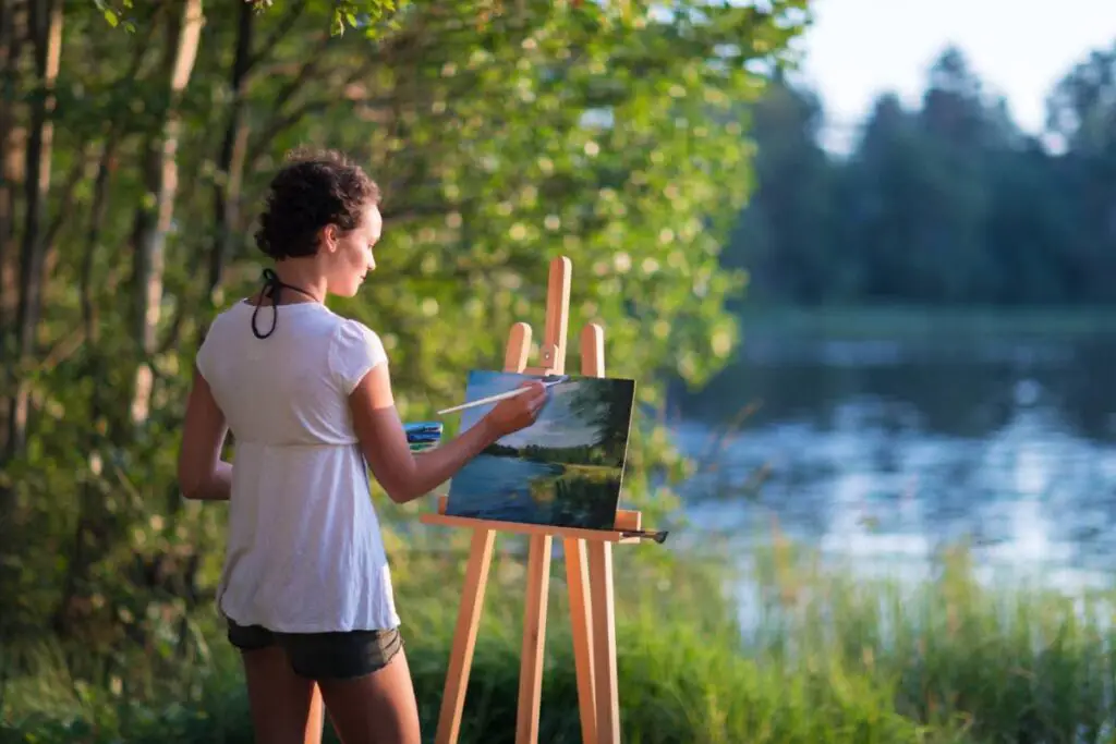 Young pretty woman artist draws paints a picture of a lake on open plain air outdoors