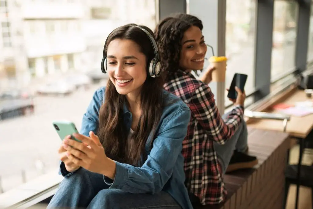 2 smiling girls sat back to back listening to music on headphones