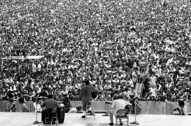 Photo of a band on stage from behind taking in the crowd they face - https://www.billboard.com/culture/events/woodstock-originally-reported-1969-8527413/