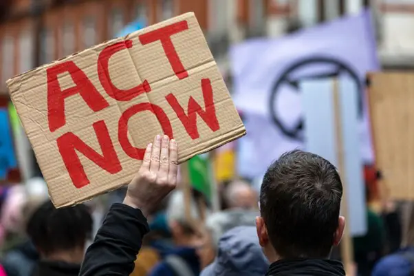 man in crowd of protesters holding cardboard sign saying ACT NOW -Source: Pexels