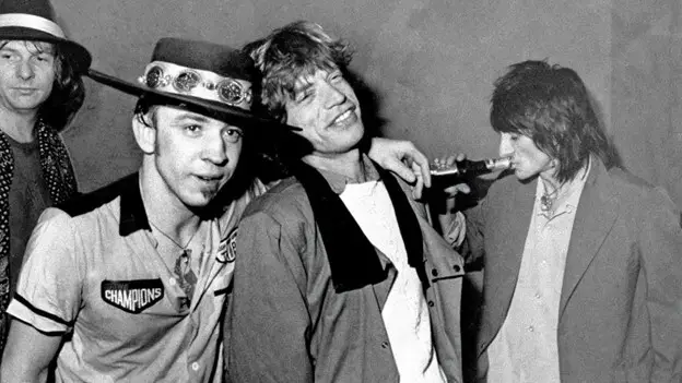 Stevie Ray Vaughan pictured with his arm around Mick Jagger. Ronnie Wood is in the background drinking from a beer bottle.