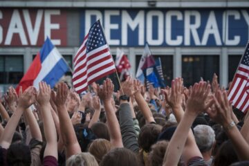 A photo of a large crowd of people with their hands raised in the air. There are multiple flags, including the American flag, being heldup by the crowd. The background contains a building with the words "Save Democracy" written in large letters.